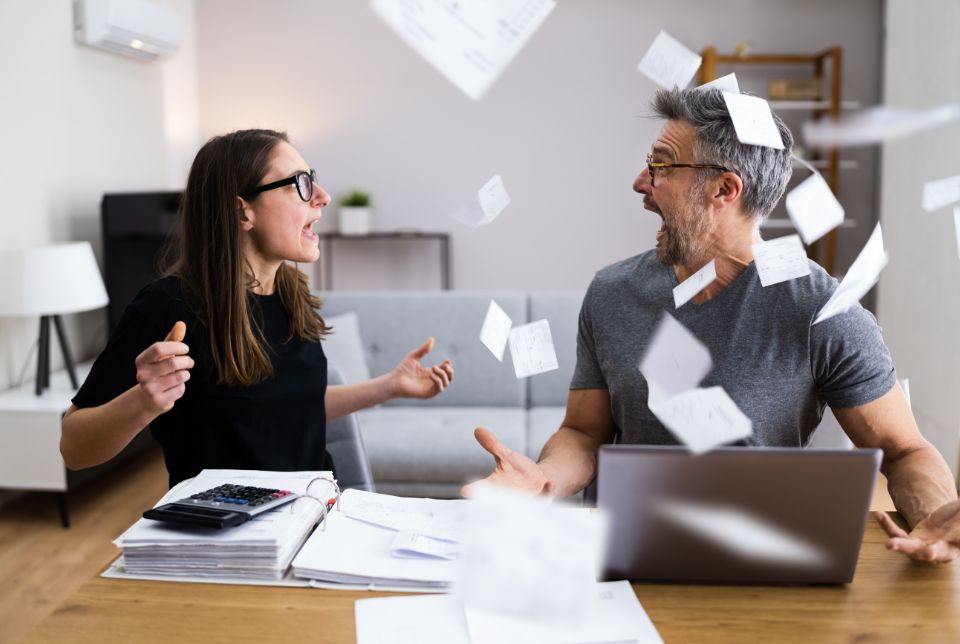 Casal discutindo e jogando papéis de contas no ar, frustrado com as dívidas antes de descobrir o Crédito com Garantia de Imóvel.